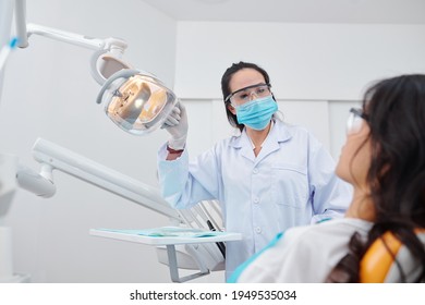 Experienced Mature Female Dentist In Protective Goggles And Medical Mask Turning On Lamp And Talking To Patient In Dental Chair