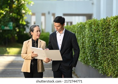 Experienced Mature Female Leader And Young Businessman Having Discussion While Standing Outdoor With Modern Office Building In Background