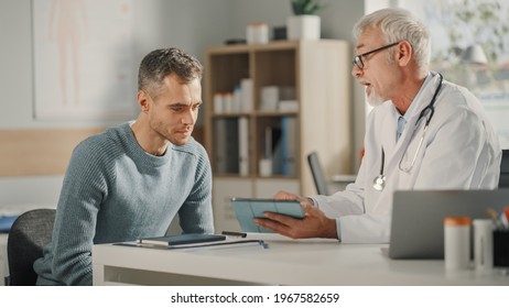 Experienced Middle Aged Family Doctor Showing Analysis Results On Tablet Computer To Male Patient During Consultation In A Health Clinic. Physician Sitting Behind A Desk In Hospital Office.