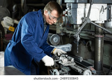 Experienced middle-aged machinist in blue work coat, protective gloves and glasses, focusing on metalworking in industrial workshop, confidently using drill press to create holes in workpiece.. - Powered by Shutterstock