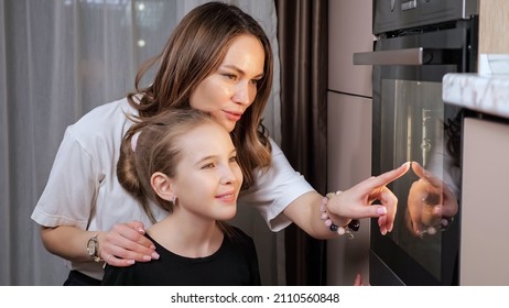 Experienced Mother Explains Teen Daughter In Black T-shirt Cooking Food Process In Stove Pointing To Glass Door In Home Kitchen Close View.