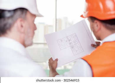 Experienced old architect and young foreman are working on plan of building. The worker is holding a blueprint. The men are looking at sketches with concentration. Focus on a paper