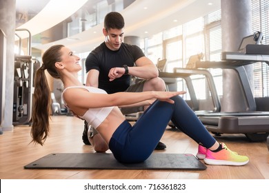 Experienced Personal Trainer Timing And Guiding Young Fit Woman During Isometric Exercise For Abdominal Muscles In A Modern Fitness Center