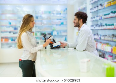 Experienced Pharmacist Counseling Female Customer In Modern Pharmacy, And Woman Pay With Credit Card