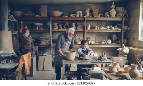 Experienced Potter Loving Prandparent Is Teaching His Young Grandchild Pottery In Small Home Workshop. Boy Is Working With Throwing Wheel, Grandpa Is Talking To Him.