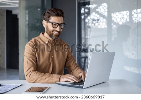 Experienced programmer inside office at workplace, senior man with beard and glasses working on laptop, businessman satisfied with achievement results.