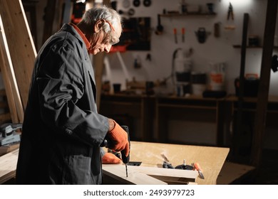 Experienced senior carpenter wearing safety glasses and gloves uses electric drill to work on wooden planks in a workshop. Concept of craftsmanship, experience, and woodworking - Powered by Shutterstock