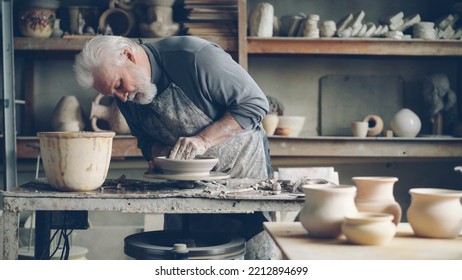 Experienced Senior Master Is Making Low Clay Bowl On A Pottery Wheel In Small Home Workshop. Ceramics Jigger, Handmade Clayware And Potter's Tools Are Visible.