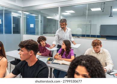 Experienced teacher distributing assignment papers to a group of diverse young adult students seated at desks in a contemporary classroom, fostering an interactive learning environment
