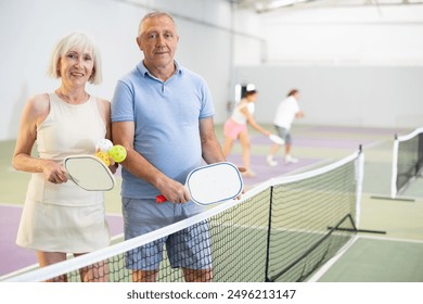 Experienced team of pickleball players, friendly smiling aged woman and man, standing on indoor court with paddles and balls in hand ready to training - Powered by Shutterstock