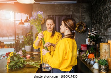 Experienced woman florist helping young employee in apron to put beautiful flowers in waze. Team of florists make bouquet in flower boutique.