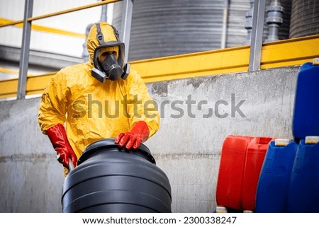 Experienced worker in protection suit and gas mask moving barrels with hydrochloric acid inside chemicals production plant.