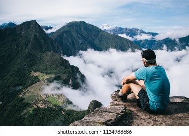 Experienced Young Man Tourist In Active Wear Sitting Rocky Hill And Relaxing While Enjoying Amazing Beauty Of Green Mountains Covered White Clouds.Traveler Admiring Natural Scenery Of Environment