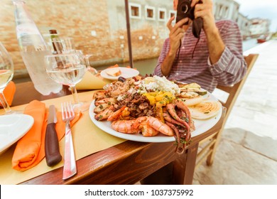 Experiential Travel Image Of Anonymous Tourist Behind The Camera Taking Photo Of Viewer, Sitting On A Table Near Canals Of Venice With Traditional Italian Seafood Platter In Foreground