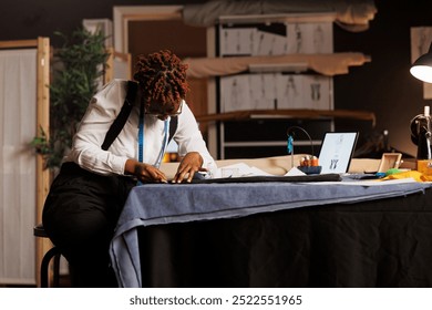 Expert focused suitmaker creating bespoken sartorial fashion collection surrounded by sewing materials and sketch drawings. African american couturier working in exquisite tailoring studio.