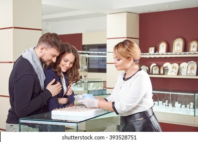 Expert Help. Female Jeweler Showing Engagement Rings To A Young Loving Couple At The Store