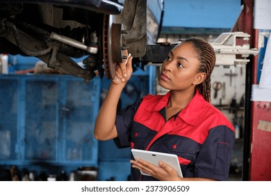 Expert young Black female automotive mechanic worker checks car's wheel brake disc and repairs inspect checklist by tablet at fix garage. Vehicle maintenance service works industry occupation jobs. - Powered by Shutterstock