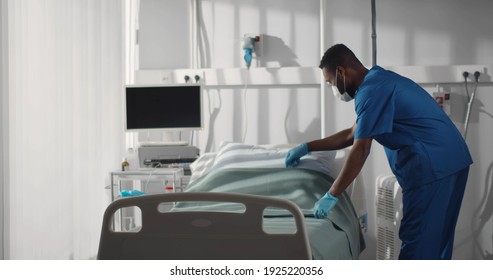 Expert Young Male Nurse In Mask Working At Hospital And Making Bed. Afro-american Man Medical Worker Cleaning Hospital Ward Preparing For New Patient