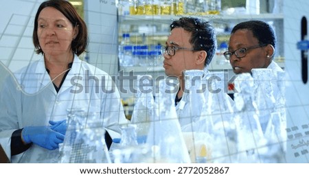 Explaining three lab staff in lab coats working at bench with flasks, shelves, blue gloves, glasses. Researchers, glassware, chemistry, collaboration, teamwork, laboratory, bright