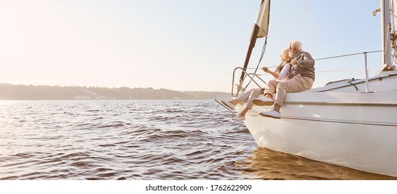 Explore dreams. A happy senior couple sitting on the side of a sail boat on a calm blue sea. Man hugging his woman while enjoying view