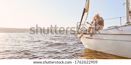 Explore dreams. A happy senior couple sitting on the side of a sail boat on a calm blue sea. Man hugging his woman while enjoying view