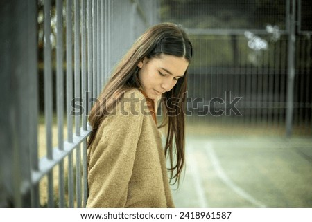 Explore the introspective beauty of a young Caucasian woman in casual clothes. With a downcast gaze, she leans on a fence, creating a captivating image