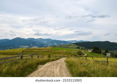 Explore the tranquil countryside with a dirt road leading through lush green fields, bordered by wooden fences, against a backdrop of majestic mountains and cloudy skies. Carpathian Mountains, Ukraine - Powered by Shutterstock