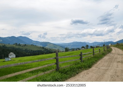 Explore the tranquil countryside with a dirt road leading through lush green fields, bordered by wooden fences, against a backdrop of majestic mountains and cloudy skies. Carpathian Mountains, Ukraine - Powered by Shutterstock