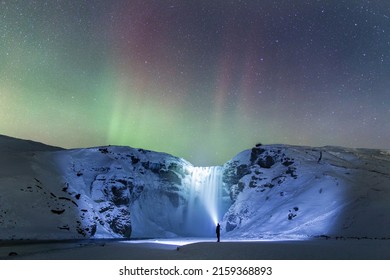 Explorer Shining Torch Light Onto A Waterfall Under The Northern Lights In Iceland.