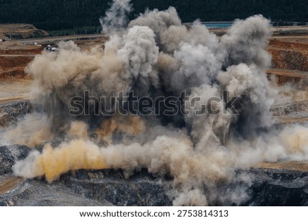 Explosive activity in large open pit gold mine with dust and smoke rising.
