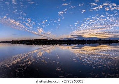 An explosive evening sunset with threads of neat cloud remnants arranged in dynamic, diagonal rows. Majestic orange patterns are symmetrically reflected in the calm river water. Tranquil spectacle.