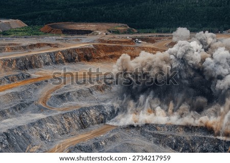 Explosive mining activity in open pit gold mine with dust clouds and heavy machinery.