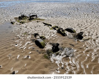 Exposed remnants of a vehicle wreck on sandy tidal flats, surrounded by rippled sand patterns under sunlight. Wheels, chassis and steering wheel still visible following years of tides.
