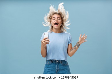 Expressing happiness with help of great tunes. Joyful amused and happy good-looking young female student jumping having fun listenign music in wireless earbuds, holding smartphone over blue wall - Powered by Shutterstock