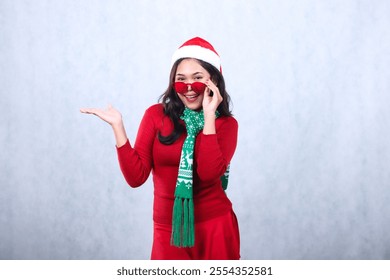 expression of asian woman wearing red sweater with Santa hat and scarf celebrating merry christmas, smiling to camera wearing red glasses presenting to the side, isolated on white background - Powered by Shutterstock