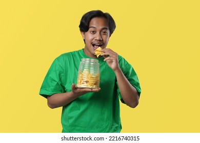 Expression happy, young asian man with green tshirt. Holds and eats a plate of chips, isolated over yellow background