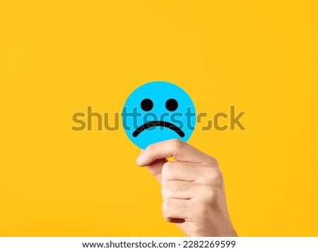Expression of negativity or customer dissatisfaction. Depression, sadness, unhappiness or joylessness. Male hand showing a blue circle with unhappy sad face on yellow background.