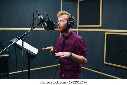 Expressive bearded man with curly ginger hair in headphones at recording studio stay opposite a microphone and performs like voice actor - Shutterstock ID 1962312676