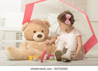 Expressive beautiful little girl having a tea party with her teddy bear, sitting under umbrella, indoor.