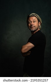 Expressive Blond Man With Medium Length Hair And A Stubble Beard In A Black T-shirt Stands With Crossed Arms In Front Of A Dark Gray Wall.