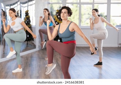 Expressive cheerful Hispanic female fitness instructor leading upbeat Zumba class for group of women in mirrored studio adorned with Christmas tree on holiday eve.. - Powered by Shutterstock