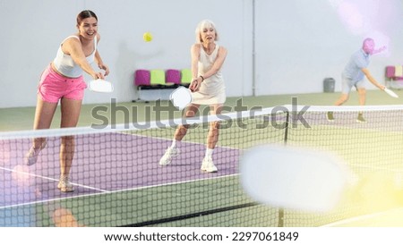 Expressive energetic girl playing doubles pickleball match in team with older woman indoors, preparing to strike and return ball. Sport and active lifestyle concept