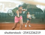 Expressive fit young girl engrossed in tennis game on clay court, hitting volley to return ball to opponent field during doubles with female partner captured in background