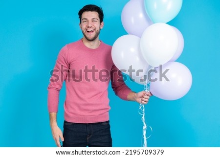 Expressive Happy Caucasian Guy Handsome Brunet Man With Bunch of Colorful Air Balloons in Pink Jumper Standing Over Blue Background. Horizontal Shot