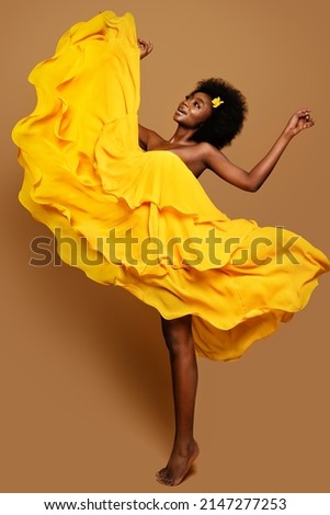 Expressive Woman dancing in Yellow Flying Dress. Happy Dark Skinned Dancer in Waving Fabric Gown. Model with Black curly Afro Hair jumping over Beige Background