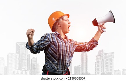 Expressive Woman In Safety Helmet Shouting Into Megaphone. Portrait Of Young Emotional Construction Worker With Wide Open Mouth On Foggy City Background. News Announcement And Advertisement.