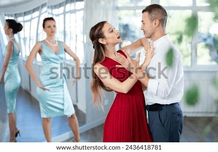 Expressive woman in vibrant red dress and man in formal wear rehearsing elegant tango in pair, participating in dance class for adults under watchful eye of female instructor