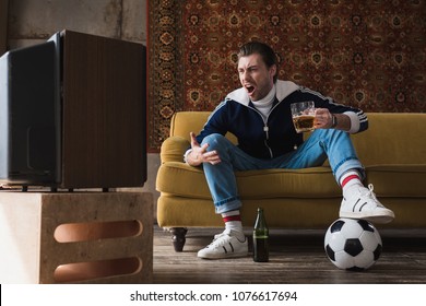 Expressive Young Man In Vintage Clothes With Ball And Mug Of Beer Watching Soccer On Old Tv And Shouting 