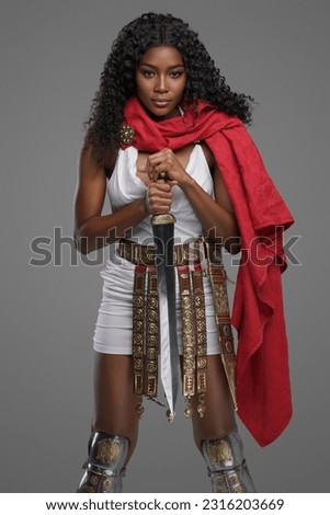 Exquisite dark skinned model in white tunic with a beautiful red cape ornate belt graves while confidently posing with gladius sword against grey background