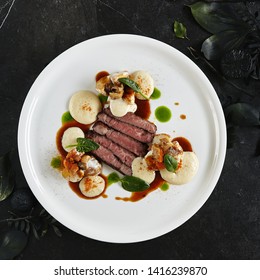 Exquisite Serving White Restaurant Plate of Black Angus Beef Fillet with Warm Potatoes in Cheese Sauce and Mushroom Espuma. Luxury Molecular Dish with Sliced Veal Meat on Black Marble Backdrop